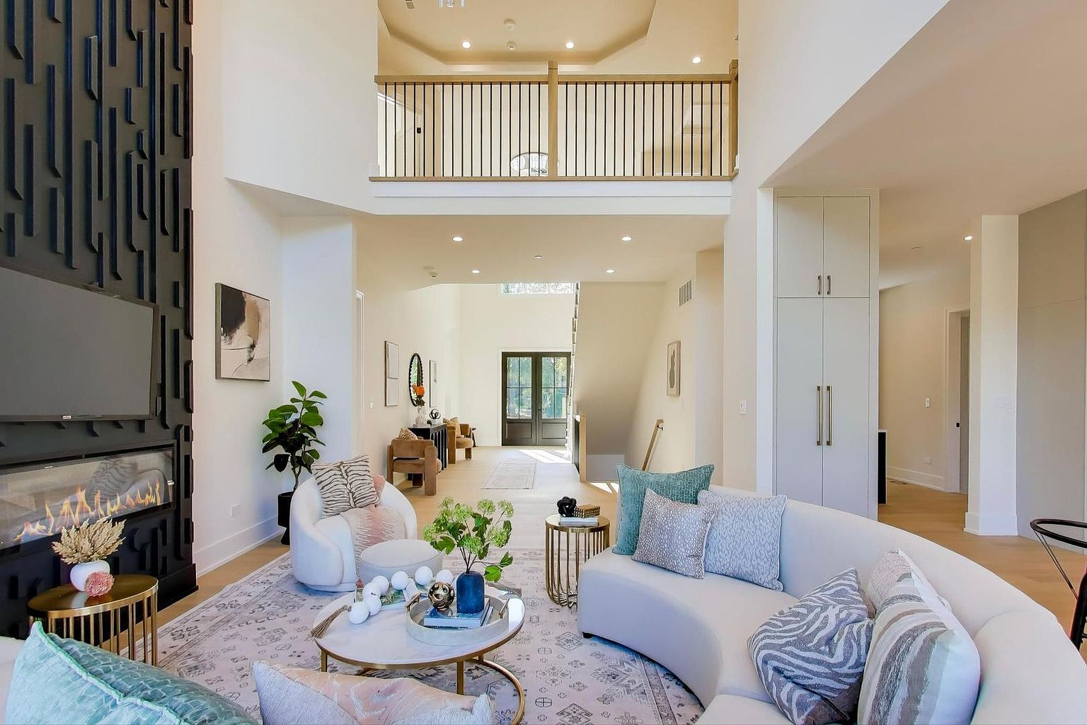 This is a spacious and luxurious living room featuring a curved sofa with decorative pillows, a modern fireplace with a large television, and a stylish coffee table. The room has high ceilings and an open floor plan, with a view of the upper level balcony. The overall impression is elegant and inviting.