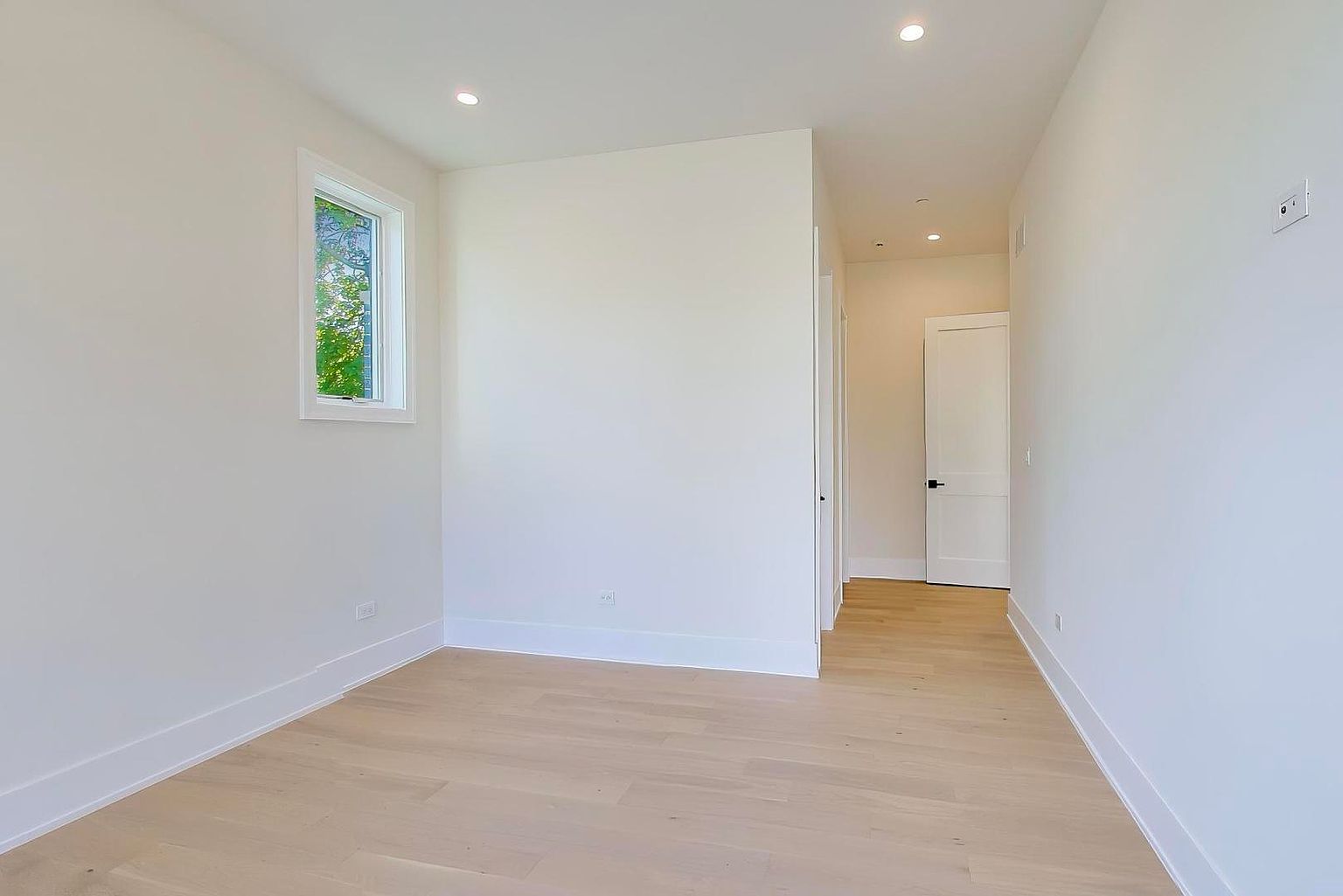 This is a bright, empty bedroom with light hardwood floors and white walls. A single window provides natural light, and the room appears clean and modern. The space is ready for personalization and furnishing, offering a blank canvas for potential buyers.