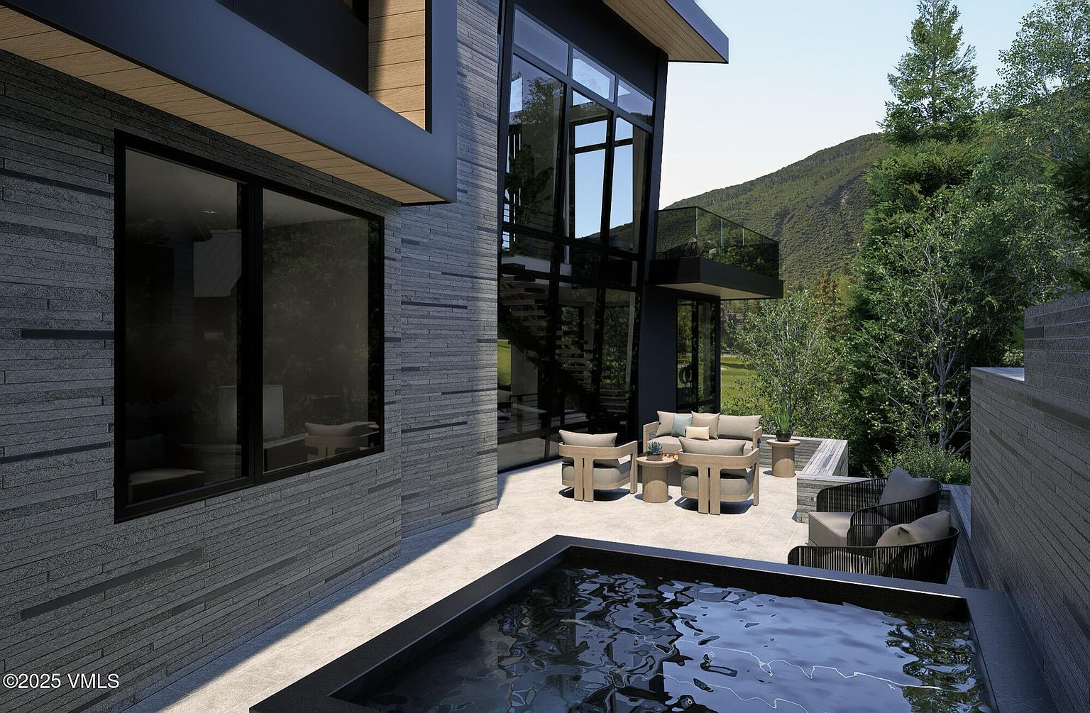 This image showcases a modern home's outdoor living space, featuring a sleek patio with contemporary furniture, including a sofa, chairs, and side tables. A dark-framed hot tub adds a luxurious touch, while the architectural design incorporates large windows and stone accents. The setting is surrounded by lush greenery and a mountain backdrop, creating a serene and upscale ambiance.