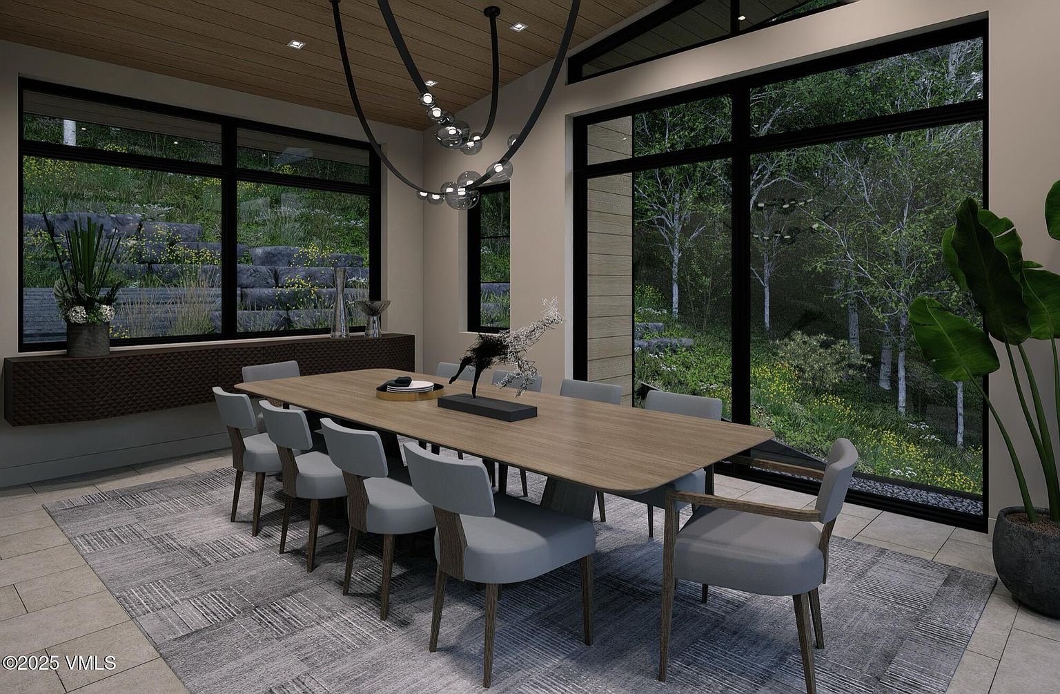 This is a dining room featuring a long wooden table surrounded by modern chairs with light gray upholstery and wooden legs. A unique, contemporary chandelier hangs above the table, and large windows offer views of lush greenery outside. The room is decorated with minimalist art and accessories, creating a sophisticated and inviting atmosphere.