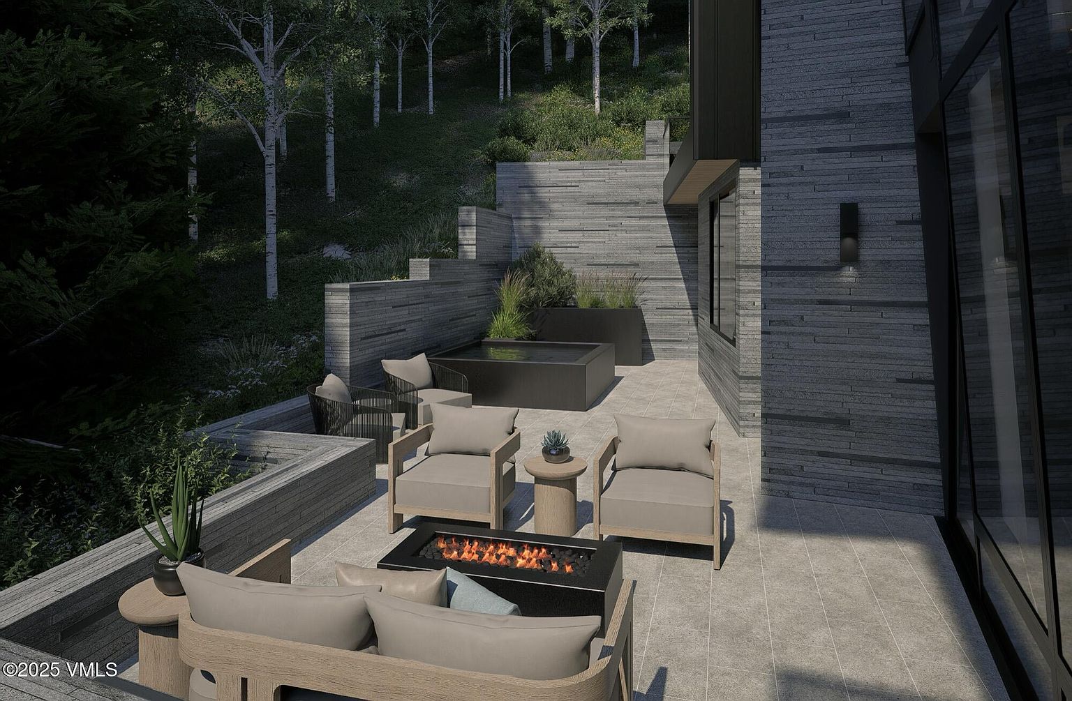 This image showcases a luxurious outdoor patio area, featuring modern furniture arranged around a sleek fire pit. The patio is constructed with gray stone tiles and is surrounded by a beautifully landscaped garden with trees and greenery. The architectural style of the adjacent building is contemporary, with gray stone cladding and large windows, creating a sophisticated and inviting outdoor living space.