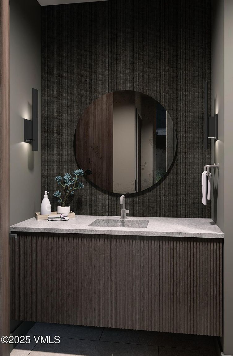 This is a modern guest bathroom featuring a dark textured wallpaper, a round mirror, and a vanity with a gray countertop and vertically ribbed wood-look cabinet. The space is illuminated by sconces on either side of the mirror, and a towel rack with white towels is visible on the right. The overall aesthetic is sleek and contemporary.