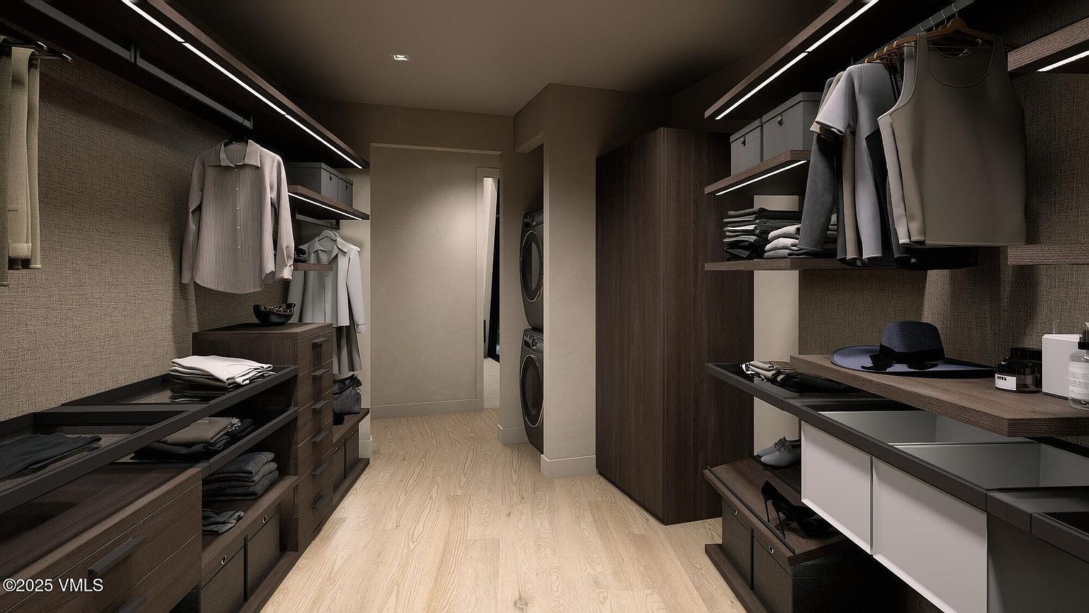 This is a well-organized walk-in closet featuring dark wood cabinetry and shelving with integrated lighting. Clothes are neatly hung and folded, showcasing ample storage space. The closet also includes a laundry area with a stacked washer and dryer, adding convenience and functionality.