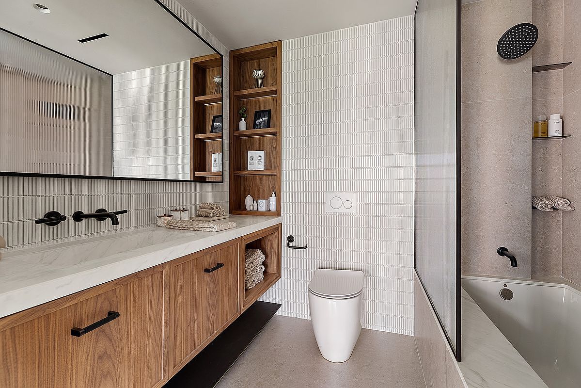 This modern, high-end bathroom features a sleek floating wood vanity with a marble countertop and integrated shelving. The space is accented by textured vertical wall tiles, a minimalist wall-mounted toilet, and a glass-enclosed shower-tub combination with matte black fixtures. The overall aesthetic is sophisticated and contemporary, emphasizing clean lines and warm, natural wood tones.