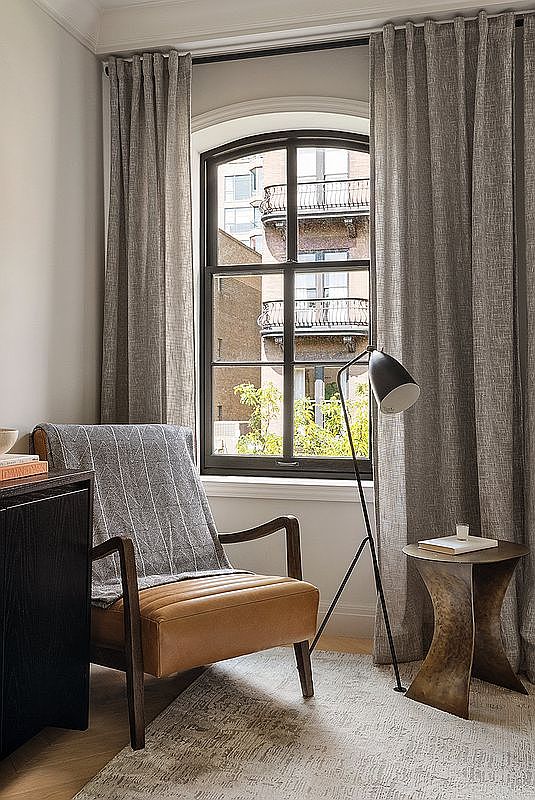 This cozy reading nook or study area features a warm tan leather armchair draped with a textured grey throw blanket, positioned next to a sleek, modern floor lamp. A unique, sculptural bronze side table sits beside the chair, while a large arched window provides a view of the neighboring building's balconies and greenery. The space is grounded by a neutral-toned area rug, creating a sophisticated and inviting atmosphere perfect for relaxation or focused work.