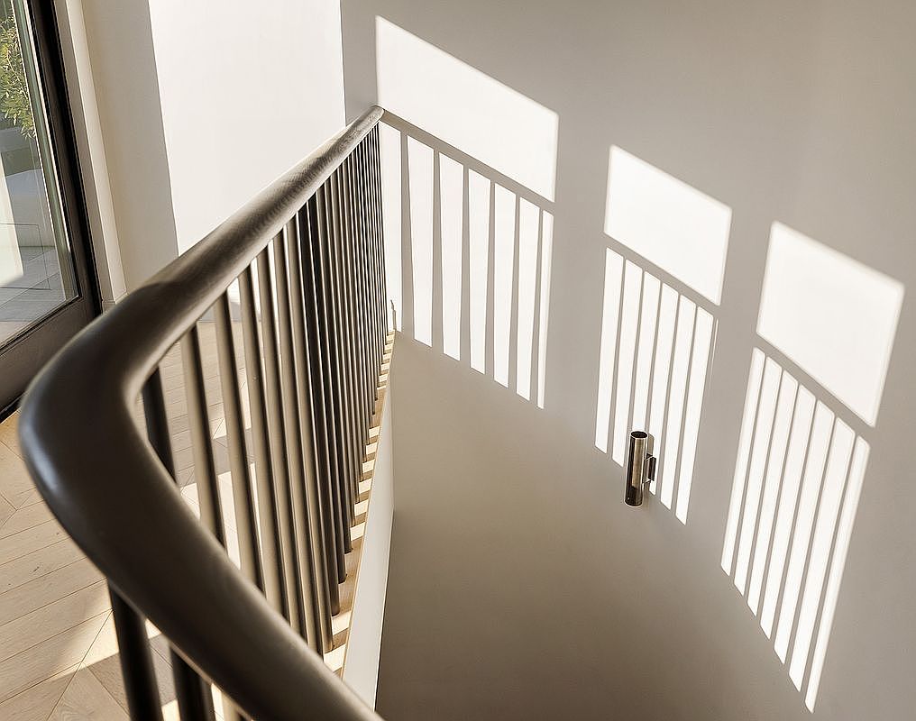 This high-angle, cinematic shot captures a sleek, modern staircase railing casting dramatic, rhythmic shadows against a minimalist light-gray wall. The interplay of natural light and geometric lines creates a sophisticated, architectural aesthetic that emphasizes clean design and open space. A single wall-mounted light fixture adds a subtle, functional detail to the otherwise stark and elegant composition.