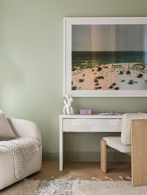 This serene home office space features a minimalist white desk paired with a modern, textured chair against a soft sage green wall. A large, framed landscape photograph of a beach scene hangs above the desk, adding a calming focal point, while a cozy, textured armchair sits to the left on a herringbone wood floor. The overall aesthetic is clean, contemporary, and tranquil, perfect for a productive yet relaxing workspace.