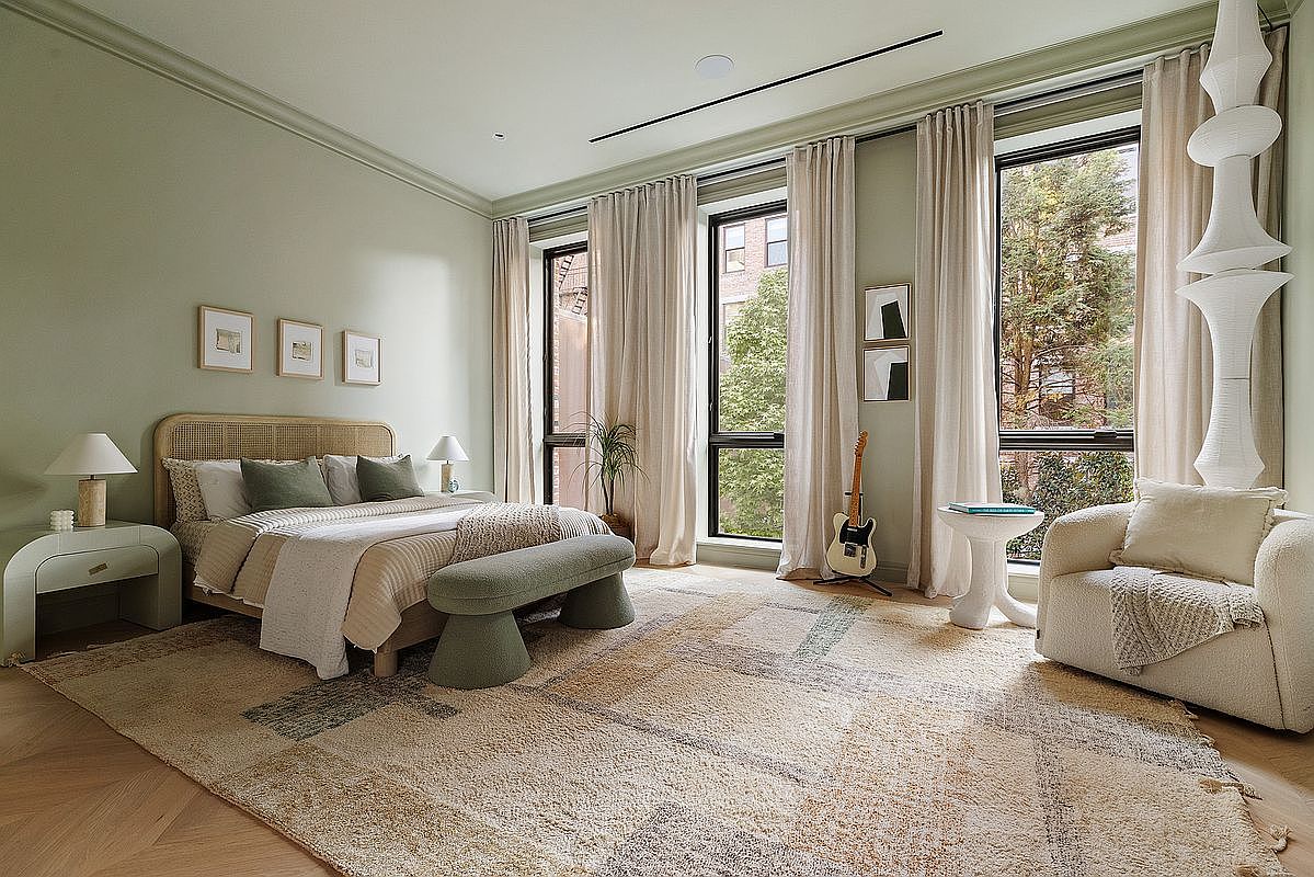 This serene primary bedroom features a sophisticated, neutral color palette with soft sage green walls and warm, textured furnishings. The room is anchored by a cane-back bed and a plush, patterned area rug, complemented by floor-to-ceiling curtains that frame large windows overlooking greenery. A sculptural floor lamp and a cozy armchair add a touch of modern elegance, creating a tranquil and inviting atmosphere.