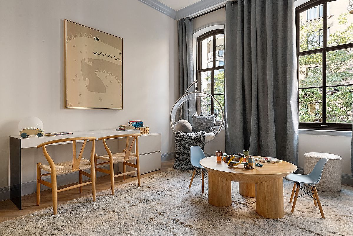 This bright and inviting playroom or study area features a modern, minimalist aesthetic with a long white desk, two wooden wishbone chairs, and a central round wooden table with matching blue chairs. A unique clear bubble chair hangs by the window, which is dressed in elegant grey curtains, while a large, textured area rug anchors the space. The room is accented by a large abstract art piece on the wall, creating a sophisticated yet playful atmosphere perfect for a family home.