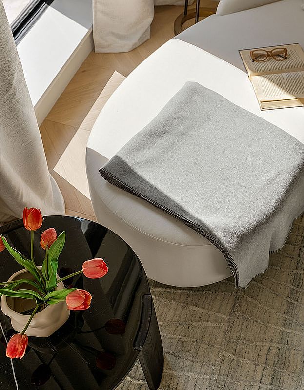 This high-angle, cinematic shot captures a serene corner of a living space, featuring a plush white ottoman draped with a soft grey throw blanket and an open book with reading glasses resting on top. A sleek, black circular side table in the foreground holds a minimalist vase filled with vibrant orange tulips, adding a pop of color to the neutral palette. The scene is bathed in soft, natural light, creating a peaceful and inviting atmosphere that suggests a sophisticated, well-curated home.