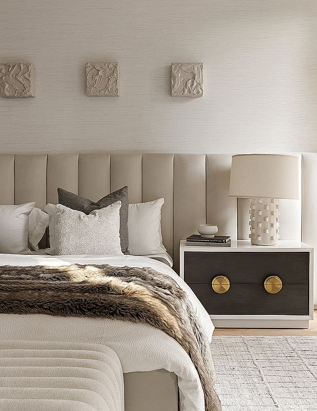 This elegant primary bedroom features a sophisticated, neutral color palette with a plush, vertical-channel upholstered headboard as the focal point. The space is accented by a dark wood nightstand with gold circular hardware, a textured lamp, and three sculptural wall art pieces, creating a serene and luxurious atmosphere. A soft, faux-fur throw blanket draped over the bed adds a layer of comfort and texture to the refined design.