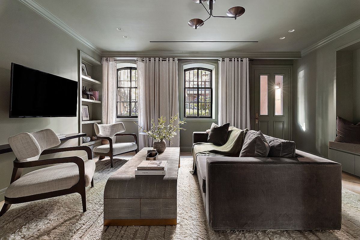 This sophisticated living room features a plush, dark velvet sofa paired with two mid-century modern armchairs and a textured, upholstered coffee table. The space is anchored by a thick, cream-colored area rug and accented by built-in shelving, elegant floor-to-ceiling curtains, and a unique modern chandelier. The room exudes a calm, refined atmosphere with its muted color palette and balanced, symmetrical layout.