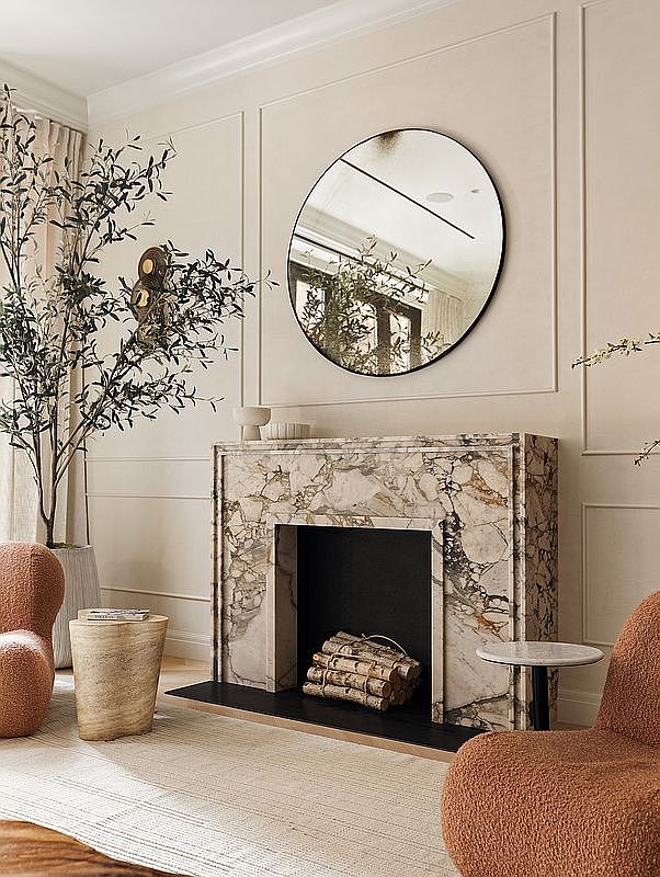This sophisticated living room features a striking marble fireplace as its focal point, complemented by a large, circular mirror and textured, warm-toned armchairs. The space is accented by a tall indoor tree and minimalist side tables, creating a serene and high-end atmosphere. The composition is captured from a straight-on perspective, emphasizing the symmetrical elegance and refined material palette of the room.