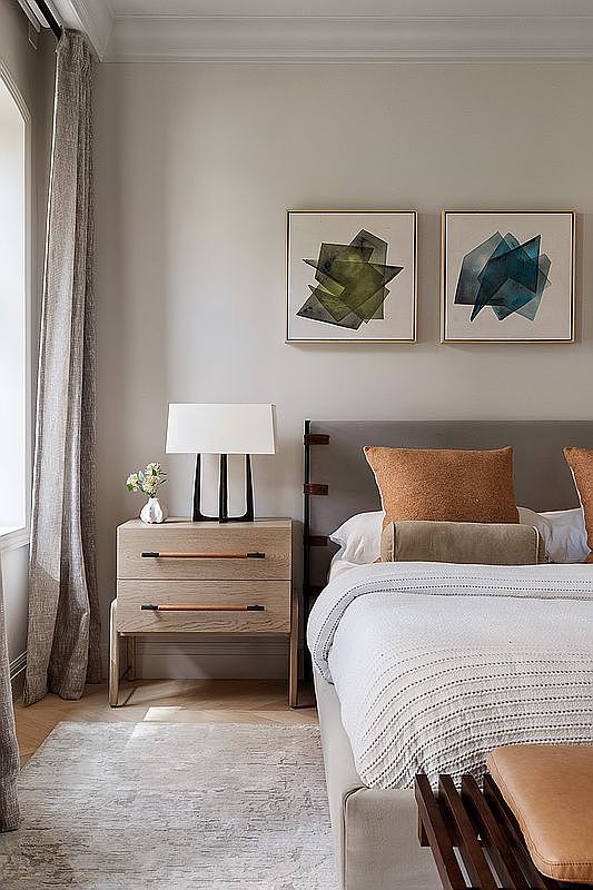 This elegant primary bedroom features a sophisticated neutral color palette, highlighted by a textured light wood nightstand with leather-accented drawer pulls and a modern table lamp. The bed is dressed in layered linens with a warm, textured throw pillow, complemented by two abstract geometric art pieces mounted above the headboard. The room exudes a serene, high-end atmosphere, enhanced by soft drapery and a plush area rug, creating a tranquil retreat.