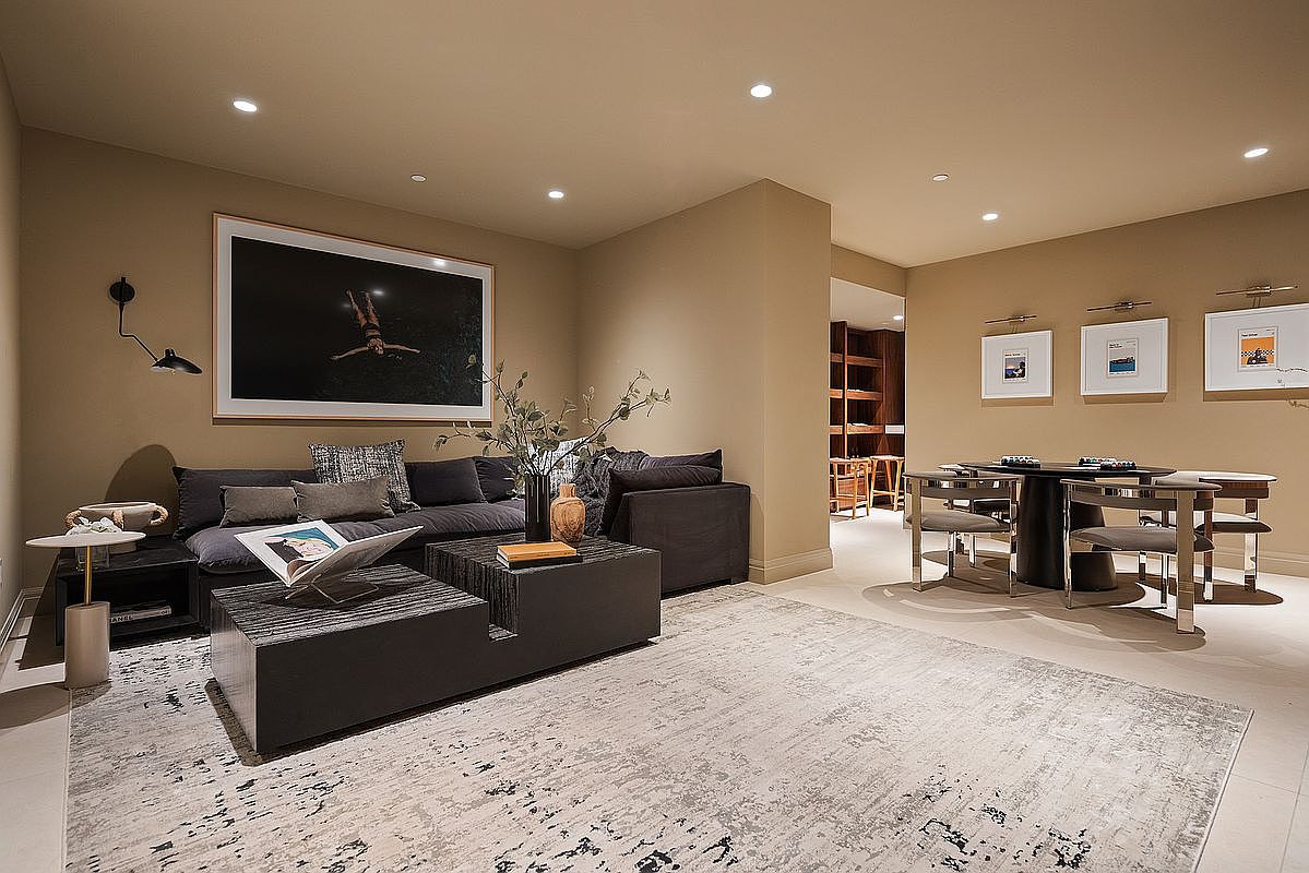 This sophisticated living room features a contemporary design with a neutral color palette, anchored by a large, dark charcoal sectional sofa and a matching tiered coffee table. A striking, oversized black-and-white photograph of a swimmer serves as a focal point above the sofa, while a nearby dining area with a round table and modern chairs adds functional elegance. The space is illuminated by recessed lighting and accented with tasteful decor, creating a warm and inviting atmosphere perfect for relaxation or entertaining.