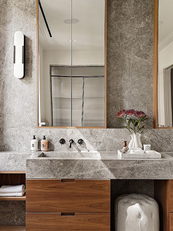 This sophisticated primary bathroom features a floating stone vanity with a sleek, integrated sink and warm wood cabinetry. The space is accented by a textured stone wall, a minimalist wall-mounted light fixture, and a decorative tray holding flowers and candles. The composition is captured from a straight-on, eye-level perspective, emphasizing the clean lines and high-end, contemporary design.