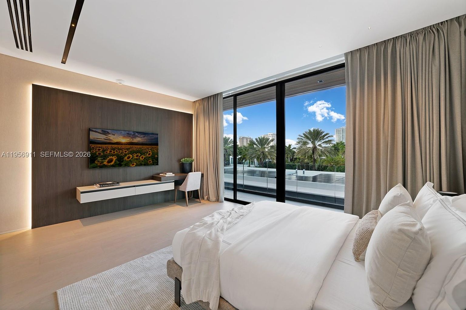 This modern primary bedroom features a sleek, dark wood accent wall with a wall-mounted television and a built-in floating desk. Large floor-to-ceiling windows provide abundant natural light and a view of the outdoor balcony and surrounding palm trees. The room is furnished with a plush bed dressed in white linens, complemented by a light-toned area rug and neutral-colored curtains.