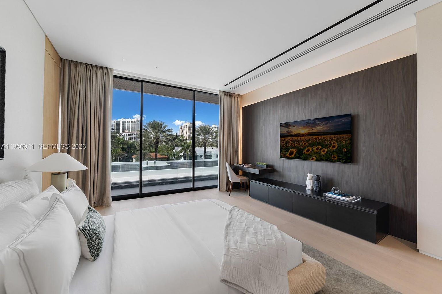 This modern primary bedroom features a minimalist aesthetic with a neutral color palette and light wood flooring. A large, dark wood-paneled wall serves as a focal point, housing a wall-mounted television above a sleek, low-profile black media console and a small integrated desk area. Floor-to-ceiling windows provide abundant natural light and offer a clear view of the outdoor balcony and surrounding cityscape.