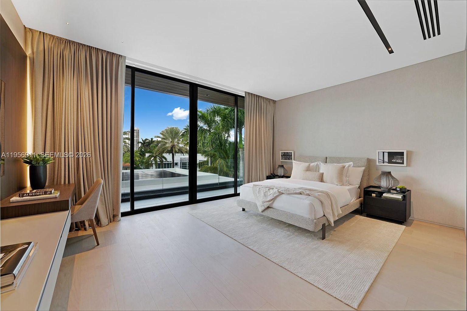 This spacious, modern primary bedroom features light wood flooring and neutral-toned walls, creating a serene and airy atmosphere. A large bed with a textured headboard sits centered in the room, flanked by dark nightstands and contemporary table lamps. Floor-to-ceiling glass sliding doors provide abundant natural light and a seamless view of the outdoor balcony and lush tropical greenery beyond.