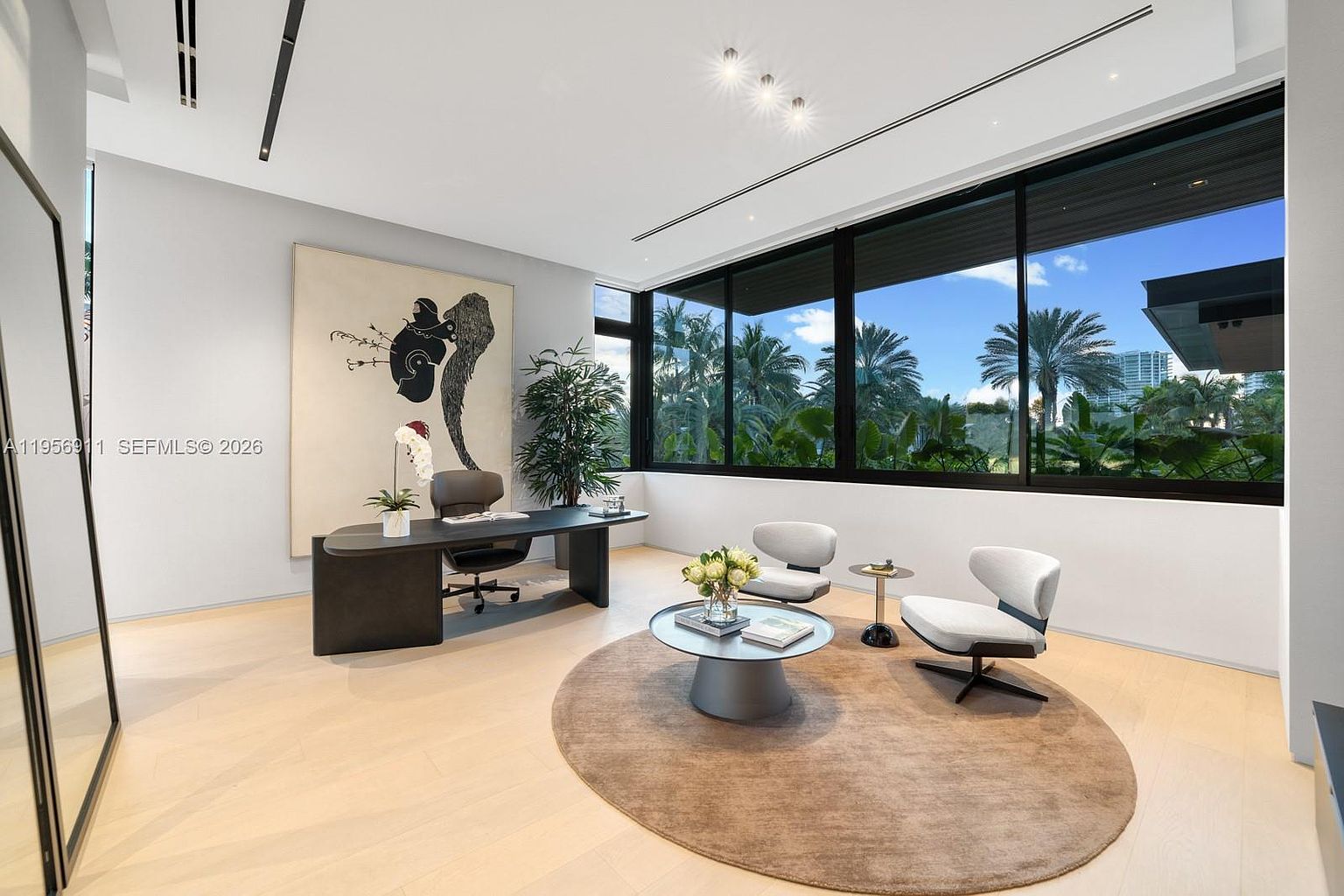 This sophisticated home office features a sleek, dark-toned desk paired with a modern ergonomic chair, set against a backdrop of light wood flooring and neutral walls. A large, abstract piece of wall art serves as a focal point, while a circular rug defines a secondary seating area with two contemporary chairs and a small side table. Large windows provide an expansive view of lush tropical greenery and palm trees, filling the room with natural light.