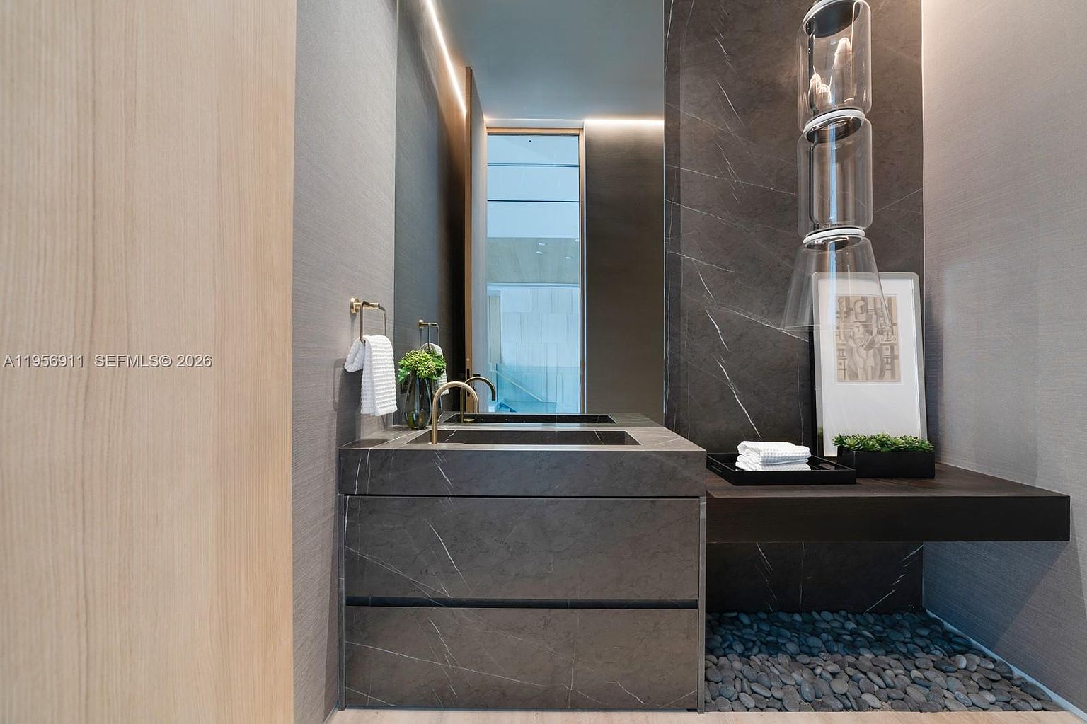 This sophisticated powder room features a dark stone vanity with a sleek integrated sink and gold-toned faucet. The space is accented by a dramatic dark stone wall, a unique stacked glass pendant light, and a decorative tray with towels. A bed of smooth river stones lines the floor beneath the floating shelf, adding a natural, spa-like texture to the modern design.