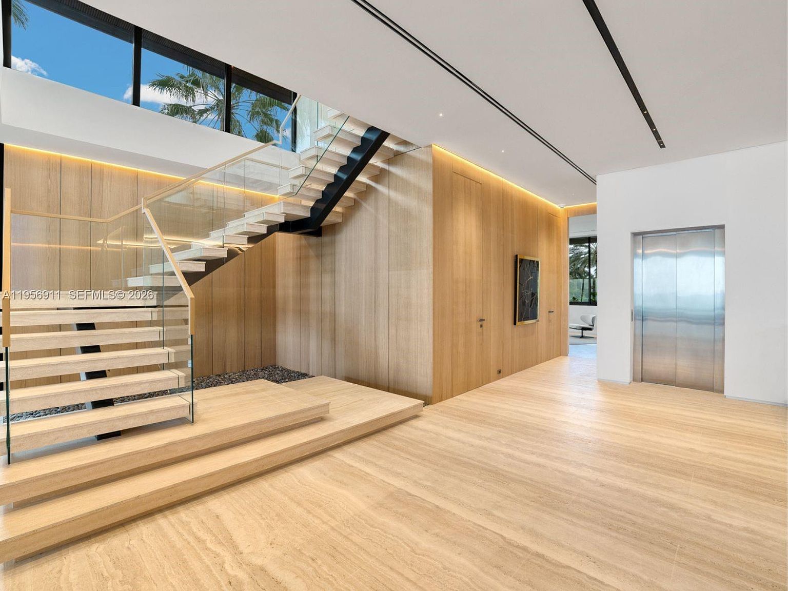 This modern, minimalist entryway features a striking floating staircase with glass railings and light wood treads. The space is defined by expansive light wood wall paneling, a polished stone floor, and a sleek stainless steel elevator door. A large window above the stairs allows natural light to flood the area, creating an airy and sophisticated atmosphere.