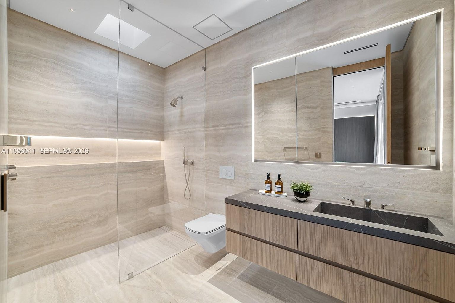 This modern bathroom features a sleek, wall-mounted vanity with light wood cabinetry and a dark stone countertop. A large, illuminated mirror spans the wall above the vanity, while a glass-enclosed walk-in shower area with neutral stone wall tiles occupies the background. The space is completed by a wall-mounted toilet and recessed lighting, creating a clean, minimalist, and high-end aesthetic.