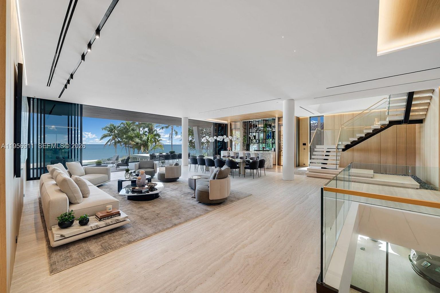 This expansive, open-concept living area features a neutral color palette with light-toned stone flooring and a large, plush sectional sofa arranged around a modern coffee table. The space is defined by floor-to-ceiling glass walls that provide an unobstructed view of the ocean and palm trees, seamlessly blending indoor and outdoor living. To the right, a floating staircase with glass railings leads to the upper level, while a sophisticated bar area and dining space are visible in the background, creating a luxurious and airy atmosphere.