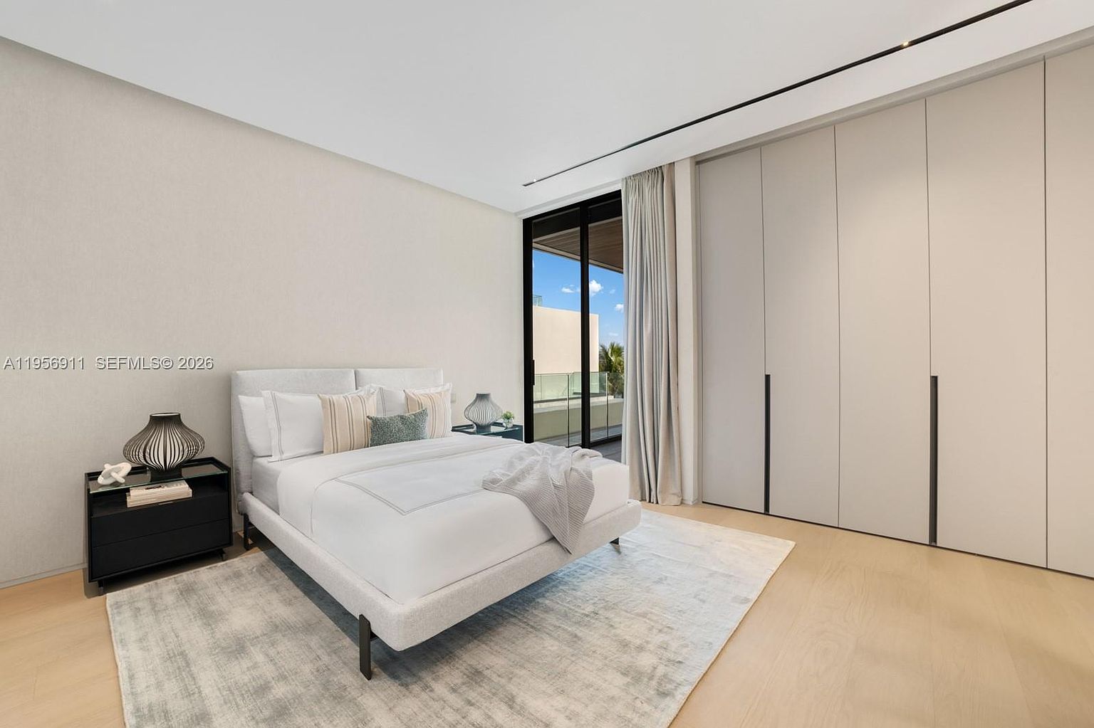 This modern bedroom features a neutral color palette with light wood flooring and a large area rug beneath a contemporary bed frame. A wall of floor-to-ceiling cabinetry provides ample storage, while a sliding glass door leads to a private balcony, allowing natural light to fill the space. The minimalist design is complemented by a sleek bedside table and a decorative lamp, creating a serene and sophisticated atmosphere.