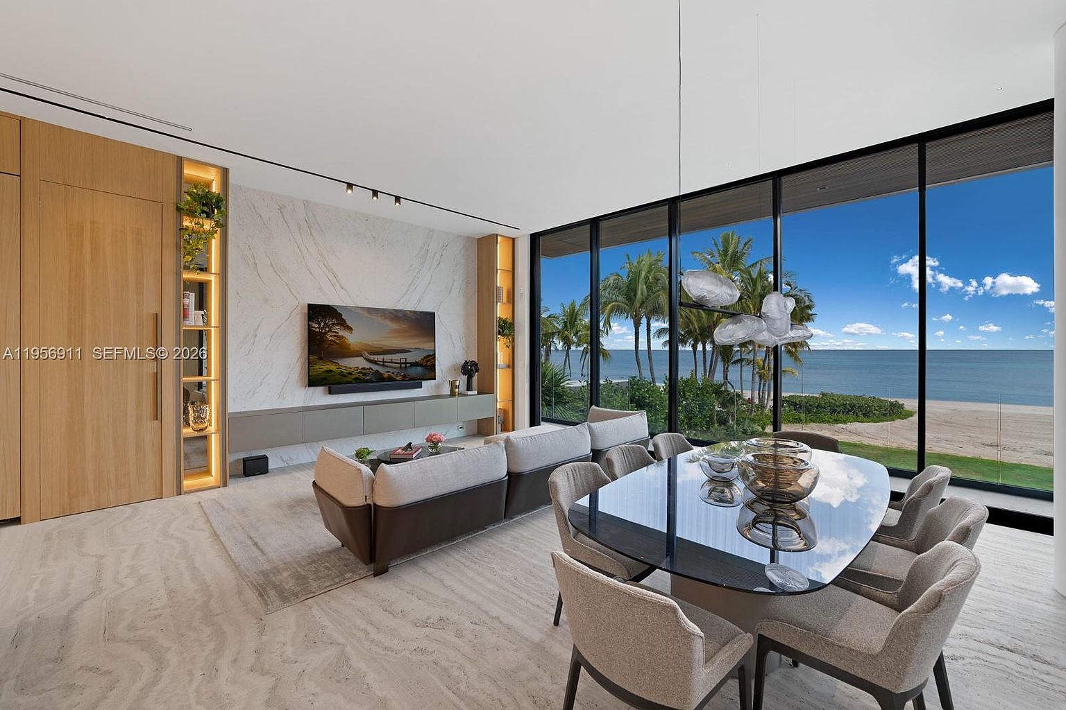 This open-concept living and dining area features a sophisticated, modern aesthetic with a neutral color palette and high-end finishes. A large, comfortable sectional sofa faces a wall with a television mounted on a light-toned stone feature wall, while the adjacent dining space is anchored by a dark, reflective table and contemporary chairs. The room is defined by expansive floor-to-ceiling windows that provide an unobstructed, panoramic view of the beach and ocean, bathing the space in natural light.