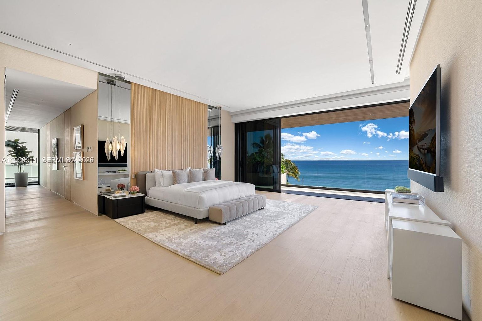 This expansive primary bedroom features a minimalist, contemporary design with light wood flooring and a textured accent wall behind the bed. A large, plush bed sits center stage, complemented by a long bench and a sleek media console, while floor-to-ceiling glass doors open to a private balcony with panoramic ocean views. The room is bathed in natural light, creating a serene and luxurious atmosphere.