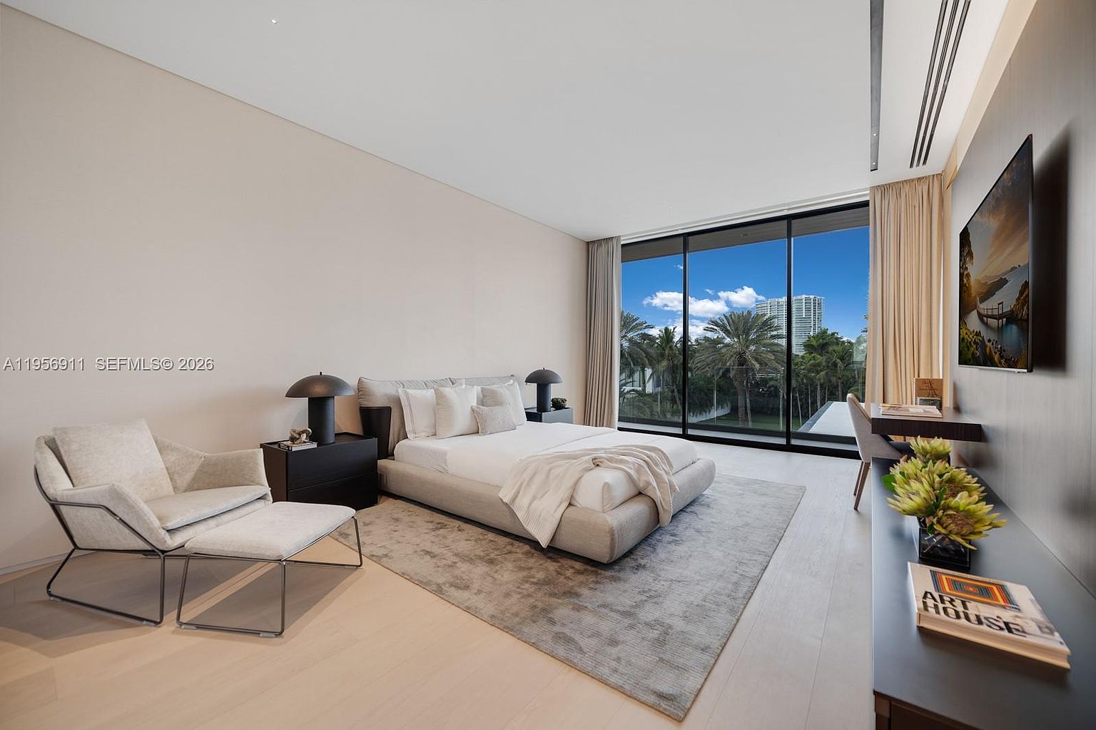 This spacious, contemporary primary bedroom features a neutral color palette and light wood flooring, creating a serene and airy atmosphere. A plush bed serves as the centerpiece, complemented by a modern lounge chair and a sleek desk area positioned near large windows that offer expansive views of the outdoors. The room is accented by minimalist decor and a wall-mounted television, emphasizing a clean, sophisticated aesthetic.