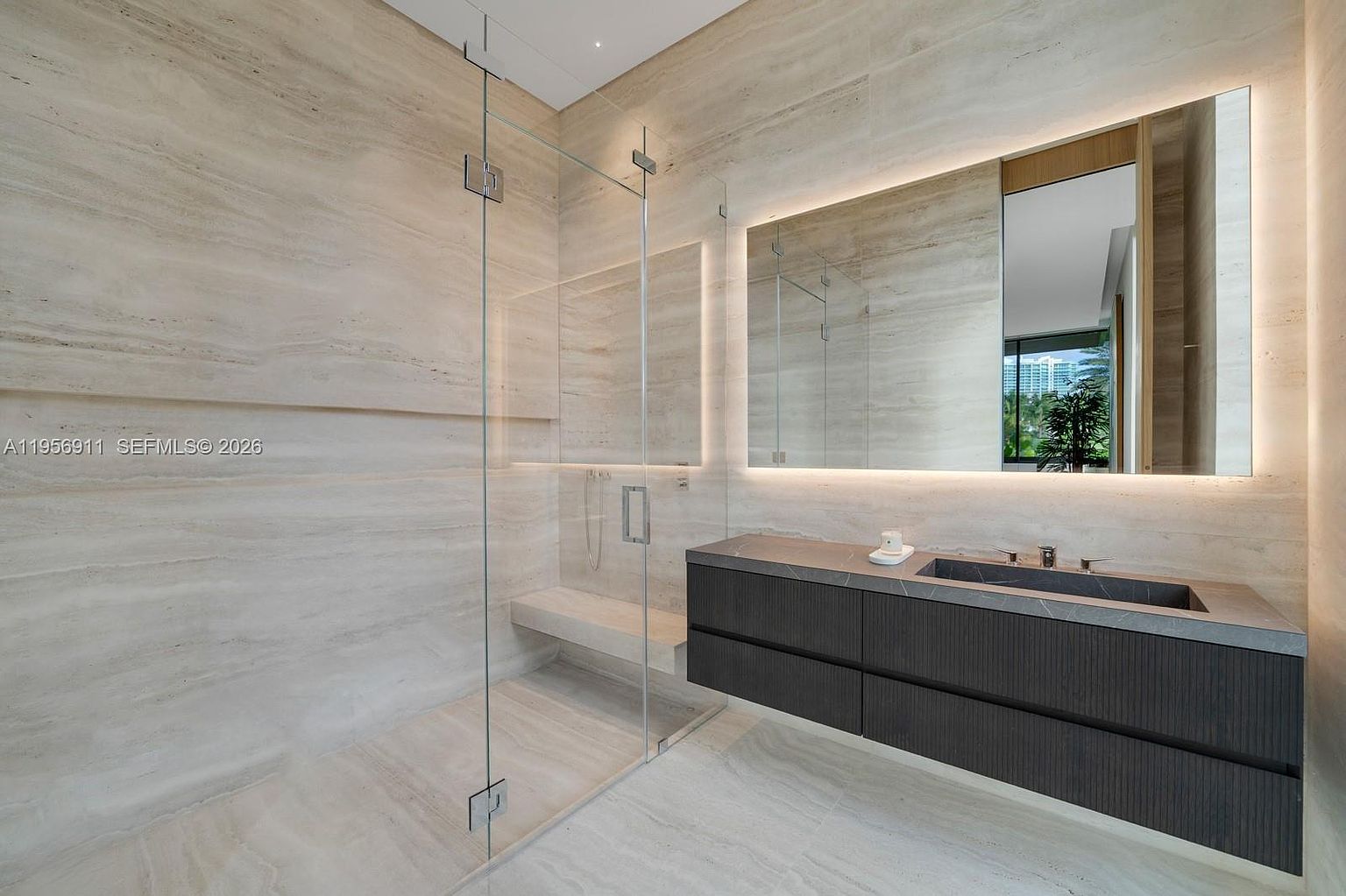 This modern primary bathroom features a sleek, dark-toned vanity with a stone countertop and an integrated sink, complemented by a large, backlit mirror. A spacious walk-in shower is enclosed by glass panels and includes a built-in stone bench, all set against walls clad in large-format, light-toned stone tiles. The minimalist design creates a sophisticated and serene atmosphere, with recessed lighting highlighting the clean lines and high-end finishes.