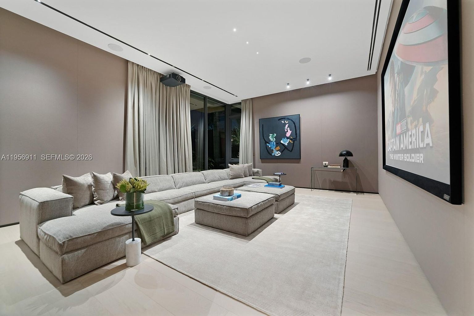 This contemporary home theater features a large, plush sectional sofa arranged around two matching ottomans on a neutral area rug. A massive projection screen displaying a movie poster dominates the right wall, while the room is finished with soft, taupe-colored walls and light-toned wood flooring. The space is accented by a modern abstract painting, a sleek console table with a lamp, and floor-to-ceiling curtains that frame the large windows.