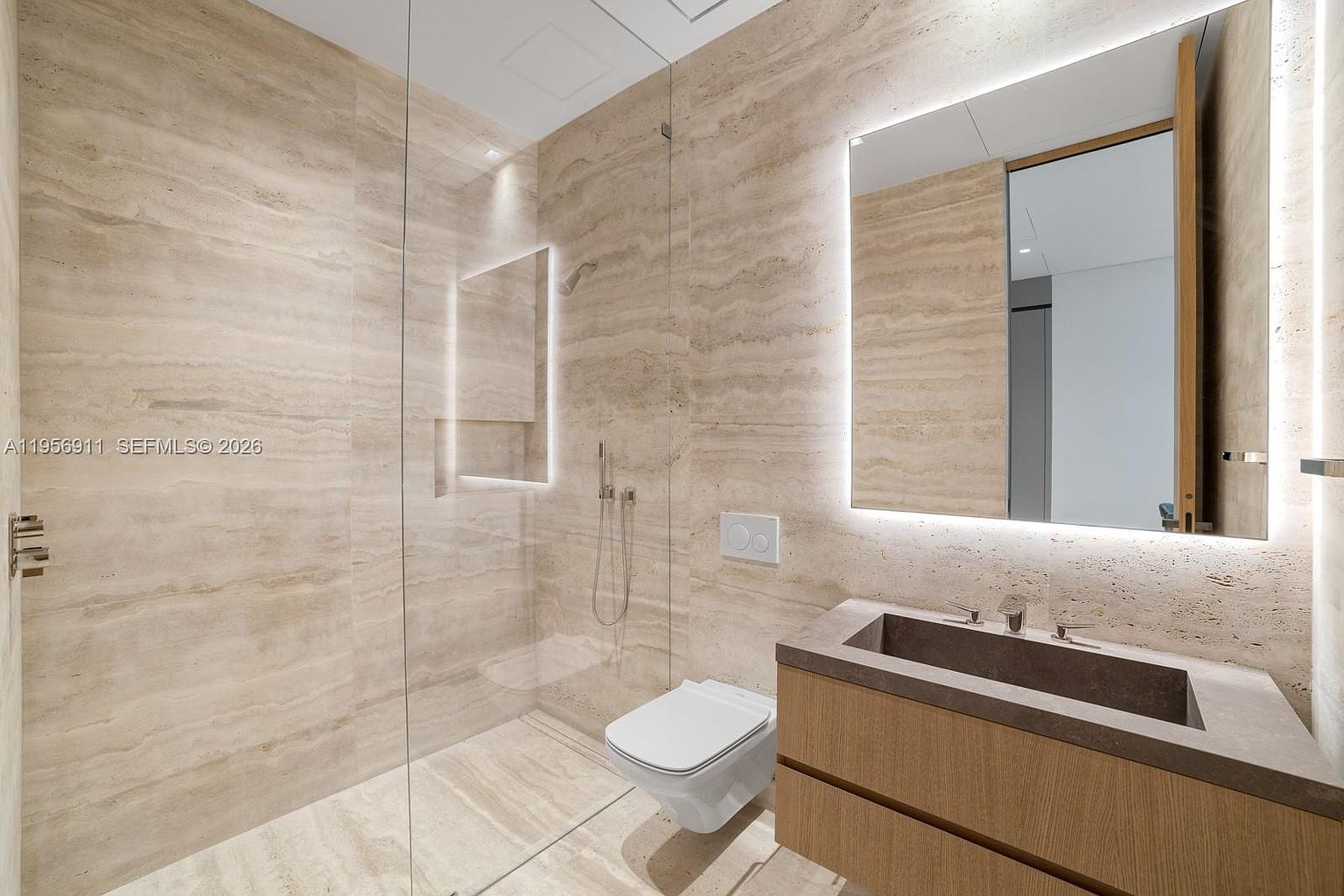 This modern bathroom features a seamless glass-enclosed shower and a floating vanity with a stone-integrated sink. The walls and floor are clad in large-format, neutral-toned stone tiles, creating a cohesive and sophisticated aesthetic. A large, backlit mirror above the vanity enhances the bright, clean atmosphere of the space.