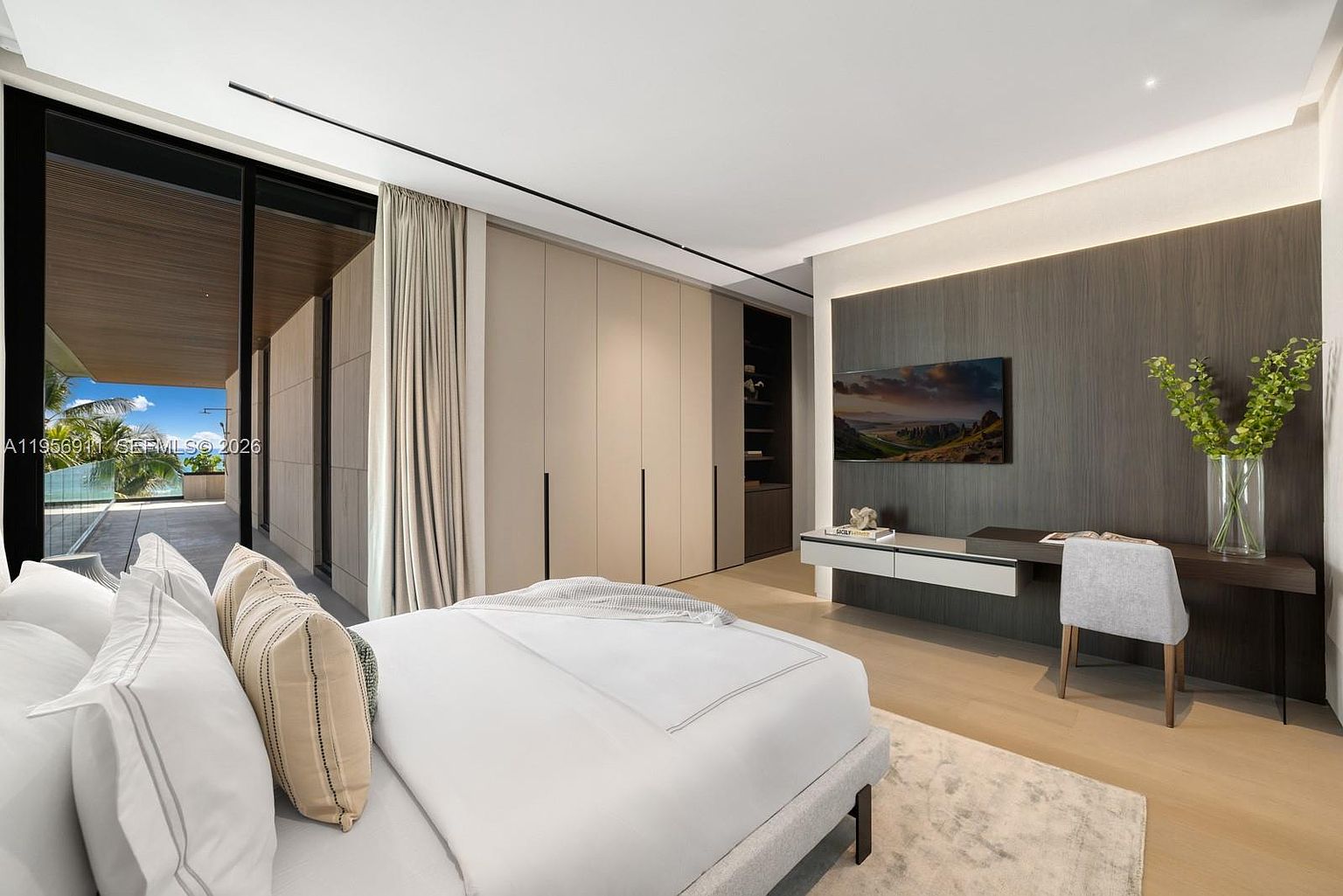 This modern primary bedroom features a minimalist aesthetic with a neutral color palette and clean lines. A large bed with plush white bedding sits in the foreground, while a dark wood-paneled wall serves as a focal point, housing a wall-mounted television and a sleek, integrated desk. Large glass sliding doors open to a private balcony, allowing for seamless indoor-outdoor living and abundant natural light.