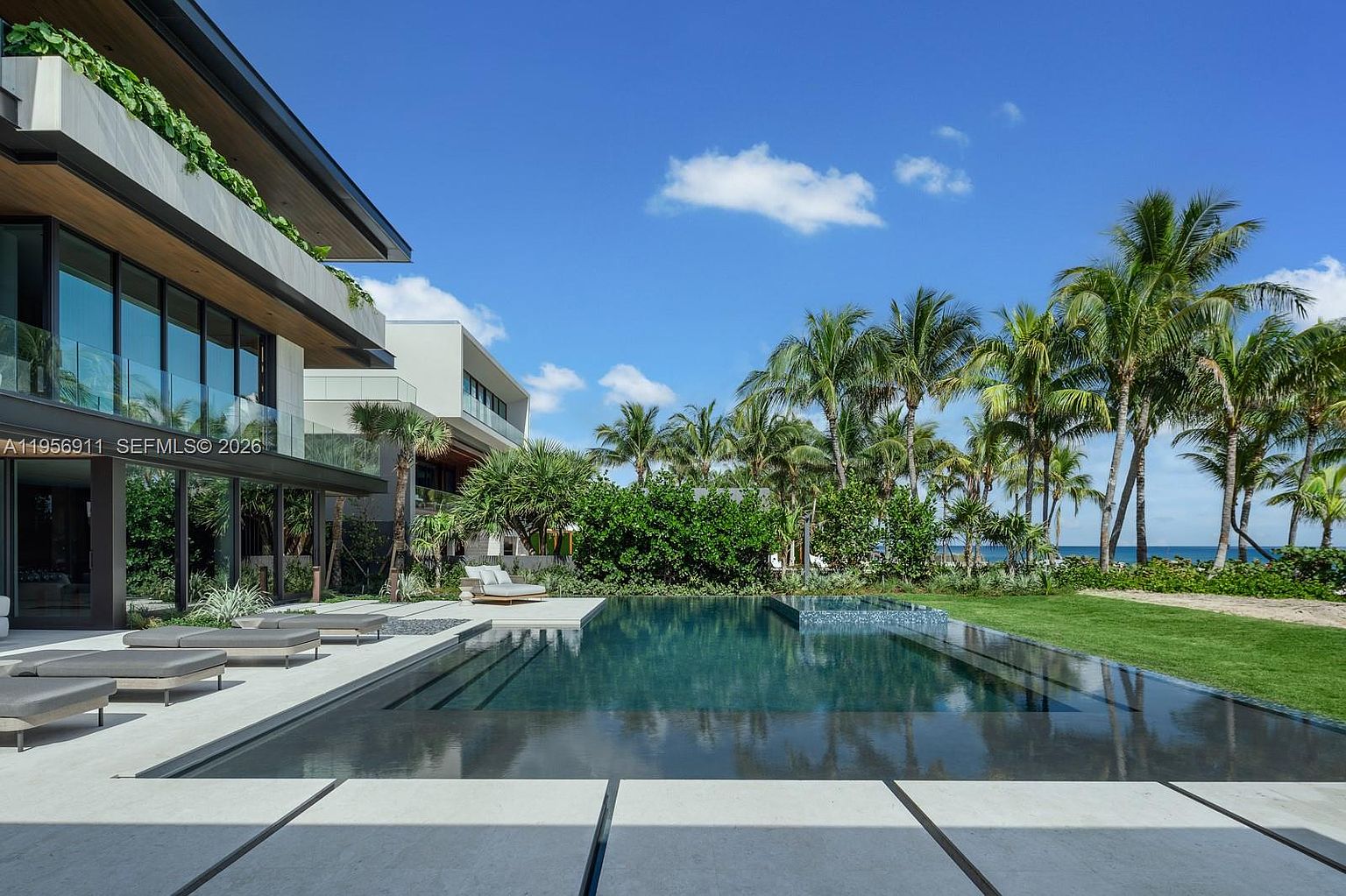 This luxurious modern residence features a sleek, dark-tiled infinity pool that seamlessly blends with the surrounding landscape. The home showcases expansive glass walls and clean architectural lines, opening onto a spacious stone patio furnished with lounge chairs. Beyond the pool, a lush green lawn transitions into a tropical garden with palm trees, offering a serene view of the ocean in the distance.