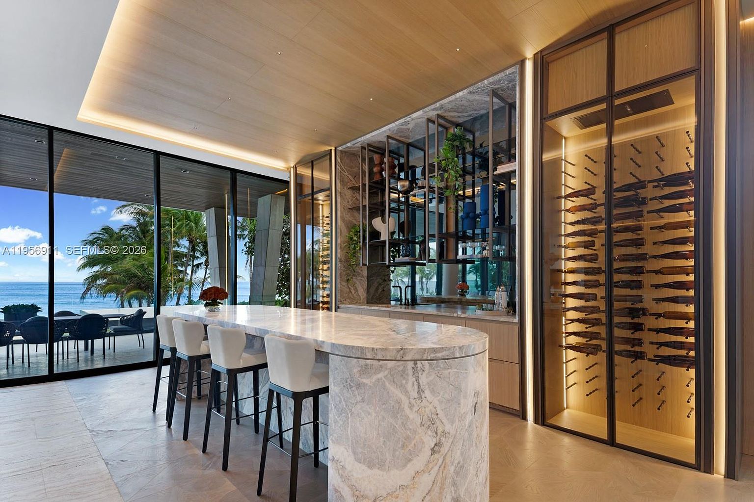 This sophisticated interior space features a large, curved stone island with bar seating, serving as a central focal point for entertaining. To the right, a climate-controlled glass wine display adds a luxurious touch, while the background showcases an open-concept layout with floor-to-ceiling glass walls that provide expansive ocean views. The room is finished with warm wood-paneled ceilings and integrated lighting, creating an elegant and seamless transition between indoor luxury and the coastal environment.