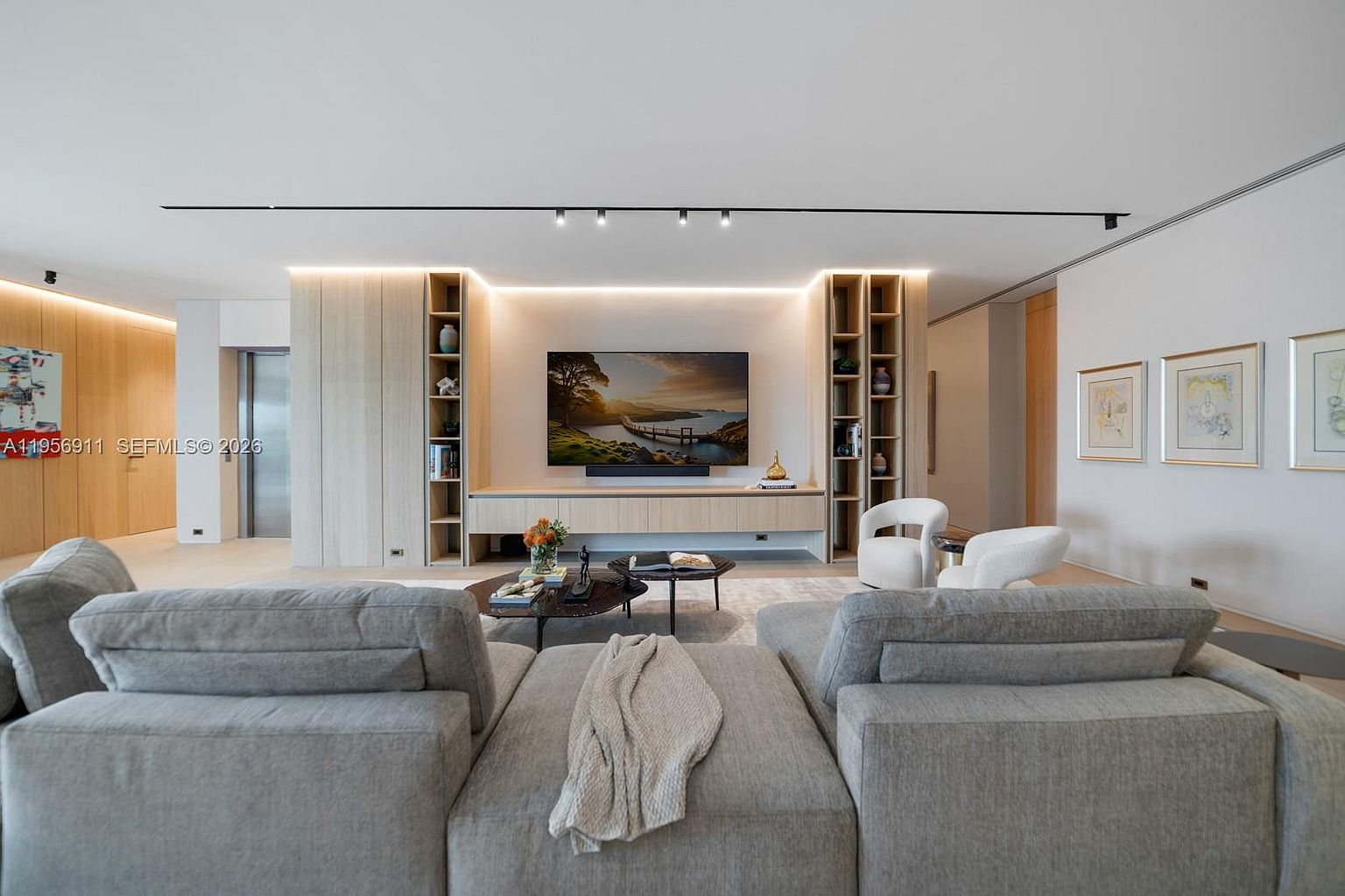 This contemporary living room features a large, light-toned sectional sofa facing a media wall with integrated shelving and a mounted television. The space is illuminated by recessed track lighting and soft ambient cove lighting, creating a warm and inviting atmosphere. The minimalist design is complemented by neutral tones, light wood cabinetry, and a pair of modern accent chairs, offering a sophisticated and comfortable living area.