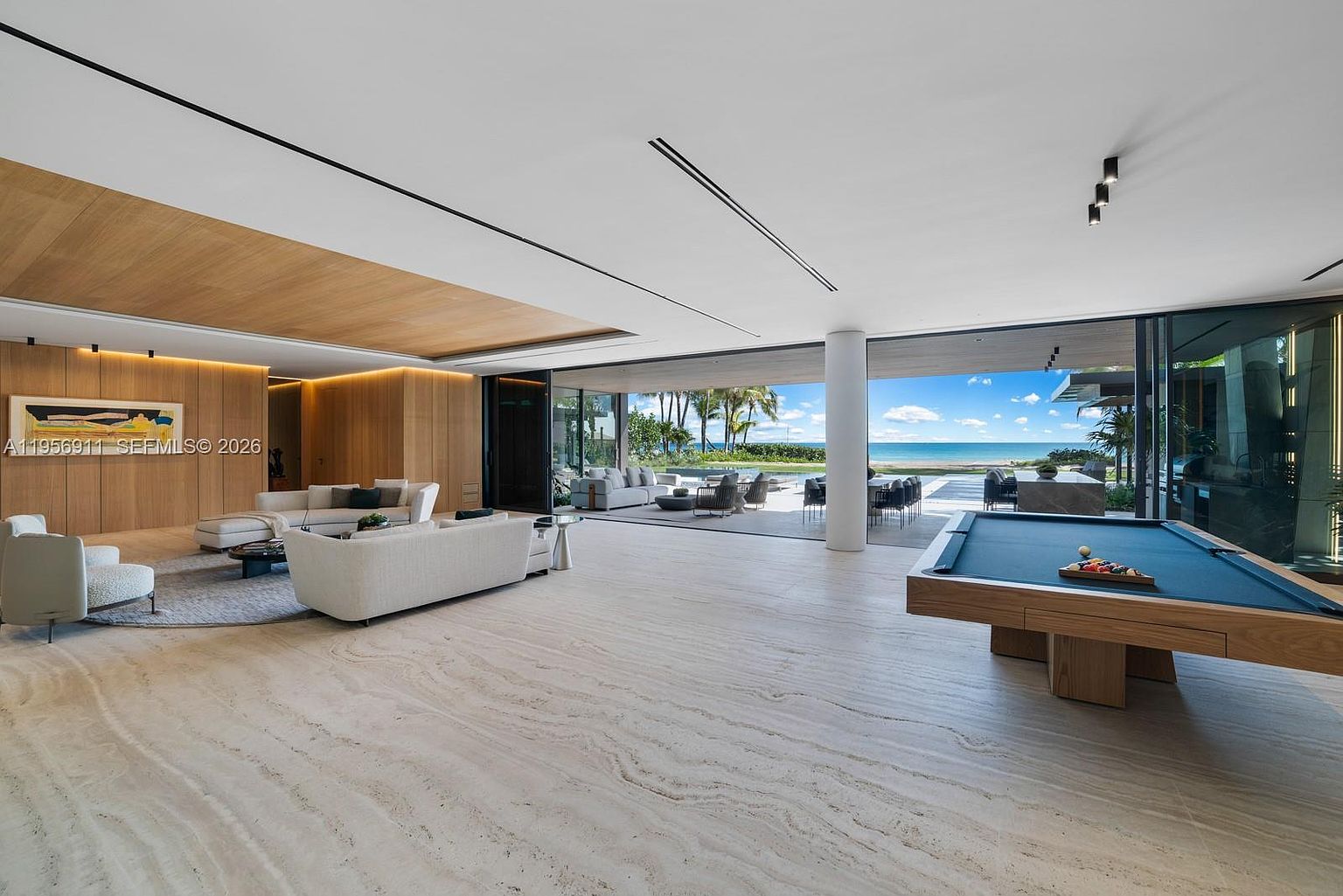 This expansive, open-concept living area features a seamless transition to an outdoor patio through large, retractable glass walls that frame a panoramic ocean view. The interior is defined by light-toned stone flooring, warm wood-paneled ceilings, and a sophisticated lounge area with plush white seating. A billiard table sits in the foreground, while the minimalist design emphasizes the connection between the luxurious indoor space and the coastal landscape.