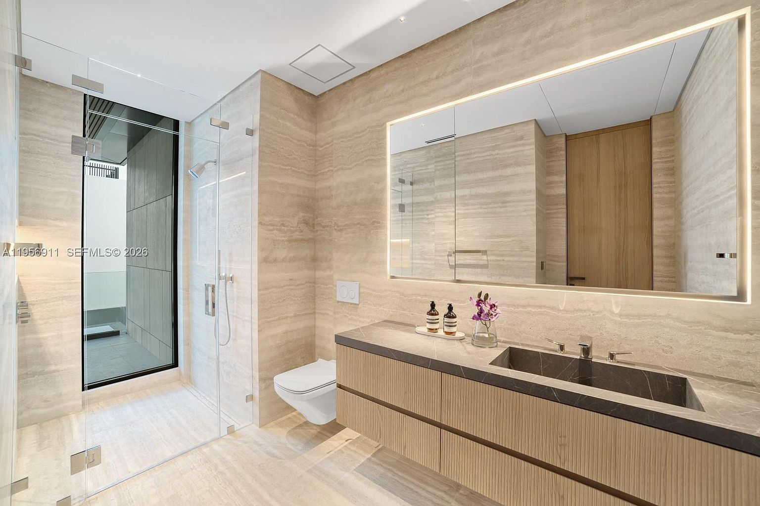This modern bathroom features a sleek, floating vanity with light wood cabinetry and a dark stone sink basin, complemented by a large, illuminated mirror. A glass-enclosed walk-in shower with neutral stone wall tiles sits adjacent to a wall-mounted toilet. The space is characterized by its clean lines, warm ambient lighting, and high-end contemporary finishes.