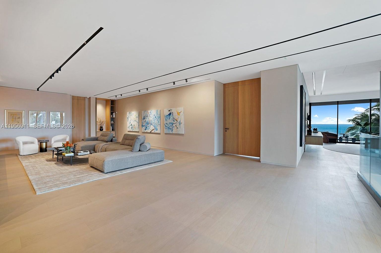 This expansive, modern living room features light wood flooring and a neutral color palette, creating an airy and sophisticated atmosphere. The space is furnished with a low-profile sectional sofa and a minimalist coffee table, complemented by abstract artwork on the wall and recessed track lighting. A large opening leads to a balcony with a stunning ocean view, emphasizing the seamless transition between indoor and outdoor living.