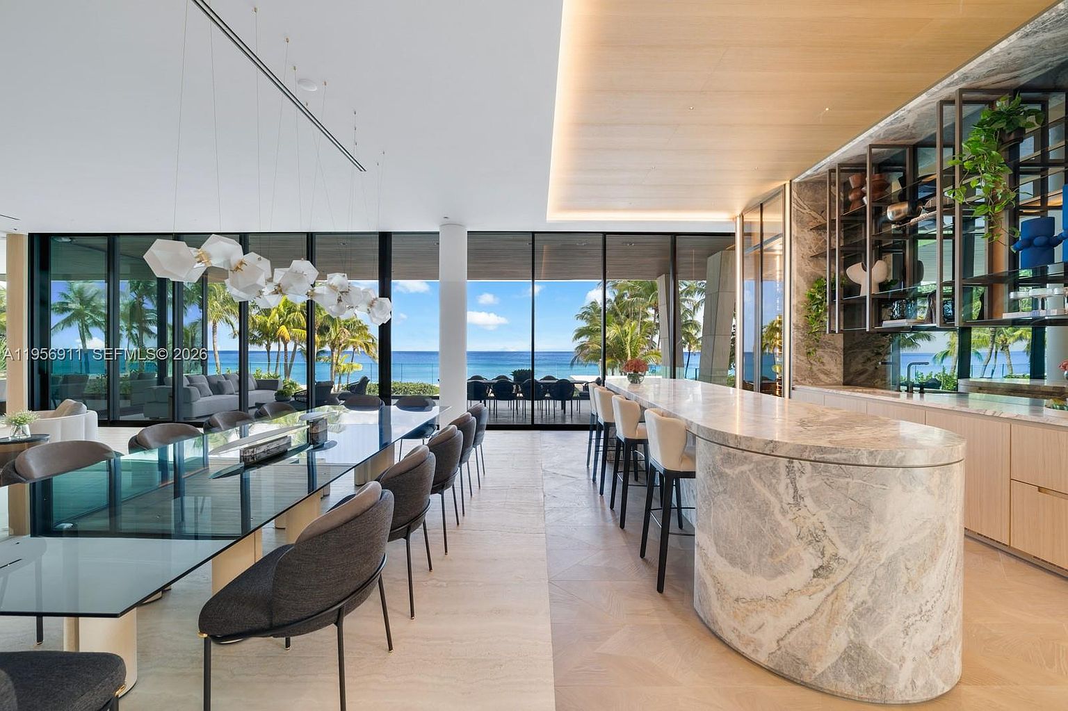 This expansive, modern open-concept space features a long, curved stone-topped kitchen island with bar seating, adjacent to a sleek glass-topped dining table. The room is defined by floor-to-ceiling windows that provide an unobstructed view of the ocean and palm trees, while a custom shelving unit with integrated lighting adds a sophisticated touch. The aesthetic is minimalist and luxurious, characterized by light-toned stone flooring and warm wood ceiling accents.