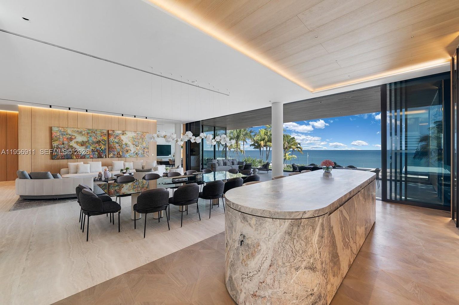 This expansive open-concept living and dining area features a large stone-topped island in the foreground and a formal dining table set for eight. The space is defined by light wood ceilings, neutral stone flooring, and a wall of floor-to-ceiling glass that frames a panoramic ocean view. A modern, sculptural light fixture hangs above the dining table, while two large abstract art pieces provide a vibrant focal point on the wood-paneled wall.