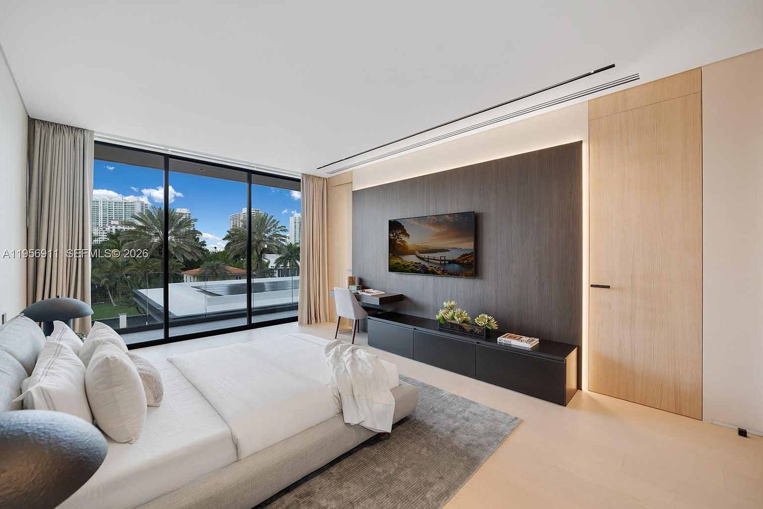 This modern primary bedroom features a minimalist aesthetic with a neutral color palette and clean lines. A large bed with light-colored bedding sits on a textured area rug, facing a dark wood-paneled accent wall that houses a wall-mounted television and a sleek, low-profile media console. Large glass sliding doors provide ample natural light and a view of the surrounding palm trees and outdoor space, while a built-in desk area adds functional workspace to the room.