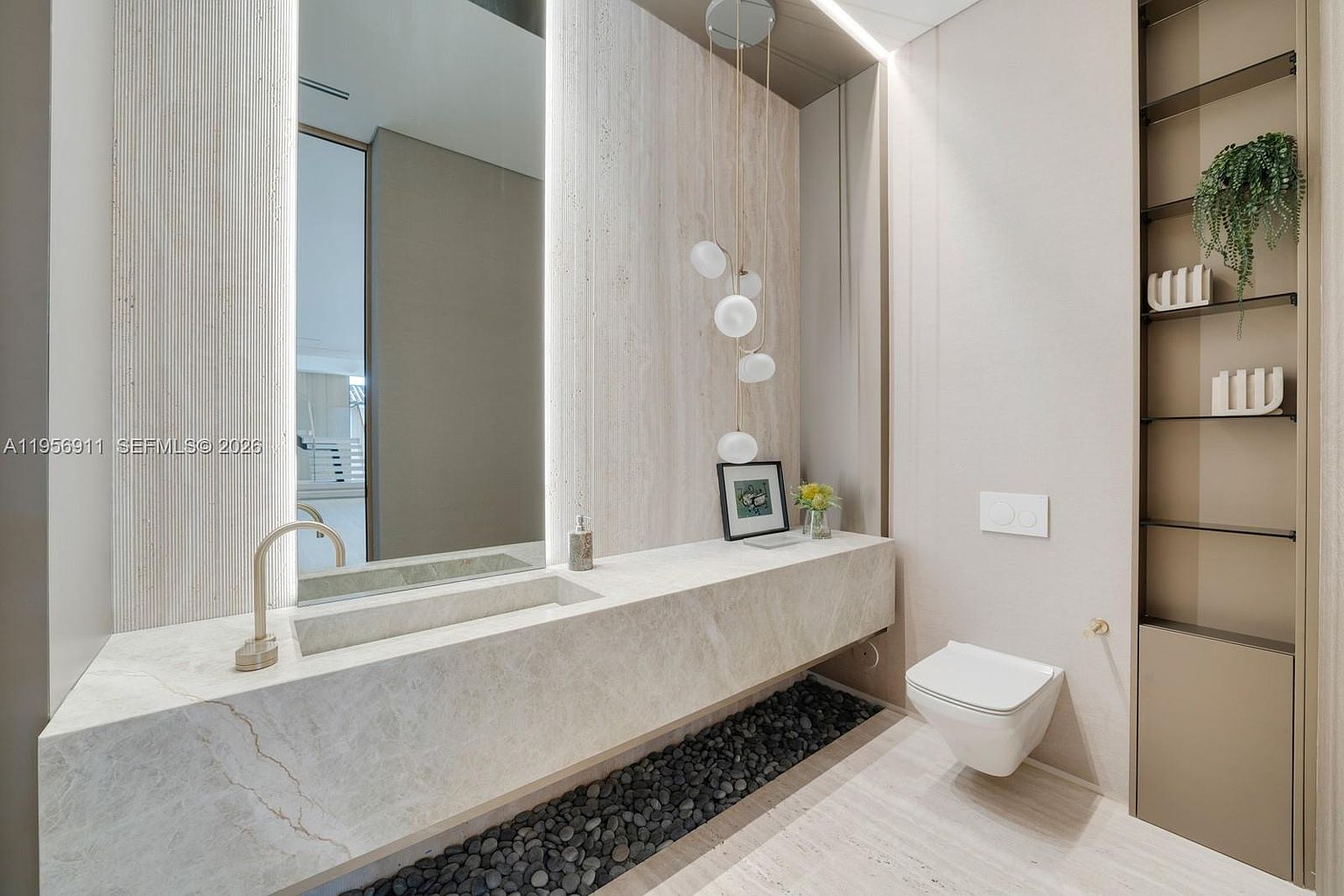 This sophisticated powder room features a floating stone vanity with an integrated sink and a sleek, wall-mounted faucet. A large, illuminated mirror is flanked by textured wall panels, while a cluster of spherical pendant lights adds a modern artistic touch. The space is accented by a decorative bed of dark river stones beneath the vanity and a built-in shelving unit, creating a serene and high-end atmosphere.