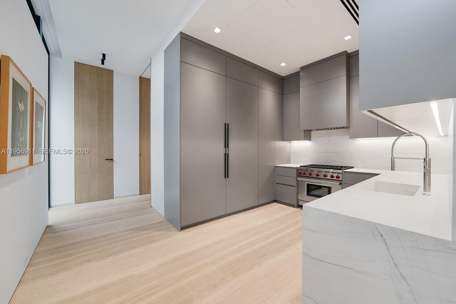 This modern, minimalist kitchen features sleek grey cabinetry, a professional-grade gas range, and a clean stone countertop. The space is characterized by light wood flooring and a bright, airy aesthetic, with a seamless transition into a hallway featuring tall wood-paneled doors. The perspective is from the kitchen island, looking toward the main cooking area and the adjacent corridor.