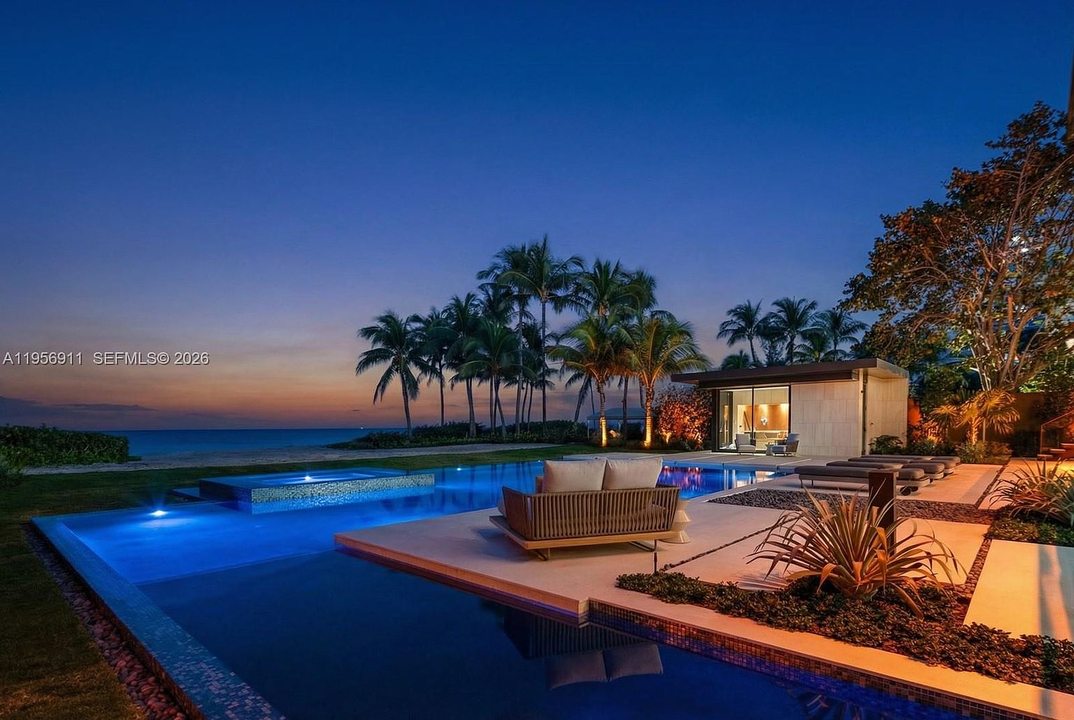 This stunning twilight view captures a luxurious infinity-edge pool and integrated spa overlooking the ocean. The scene features a contemporary outdoor lounge area with modern furniture, set against a backdrop of swaying palm trees and a vibrant sunset sky. A sleek, glass-walled cabana structure sits adjacent to the water, creating a sophisticated and serene atmosphere perfect for high-end coastal living.