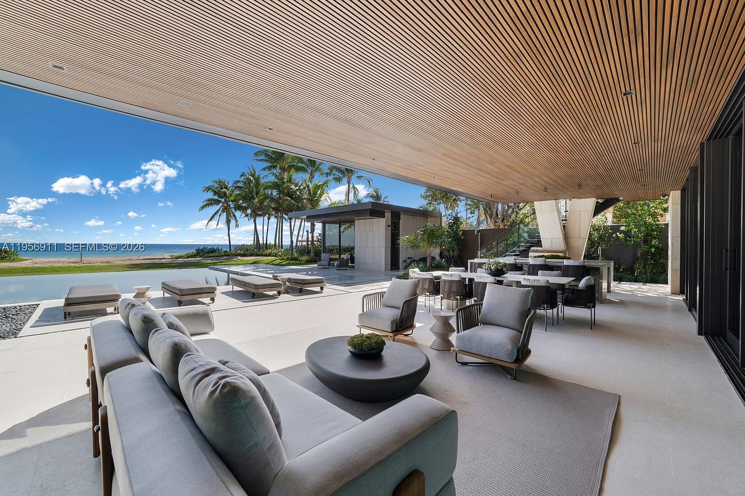 This expansive, covered outdoor living space features a seamless transition to a modern pool area overlooking the ocean. The area is furnished with a large sectional sofa, a central coffee table, and a dining set, all sheltered by a slatted wood ceiling. The open-air design emphasizes a luxurious, resort-style atmosphere with unobstructed views of the beach and palm trees.
