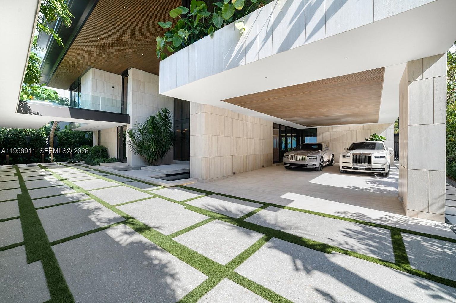 This modern luxury residence features a spacious, covered carport area capable of housing multiple high-end vehicles. The architectural design emphasizes clean lines, utilizing light-toned stone cladding and warm wood-paneled ceilings, complemented by a geometric driveway paved with large concrete pavers and integrated grass strips. The perspective captures the grand scale of the entryway and the seamless transition between the indoor and outdoor living spaces.